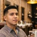 Side profile of a young Latino man with a modern low taper fade haircut, standing in a barbershop.