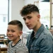 A wide shot of a young boy with a neat low fade standing next to a teenage boy with a stylish high fade in a barbershop setting.